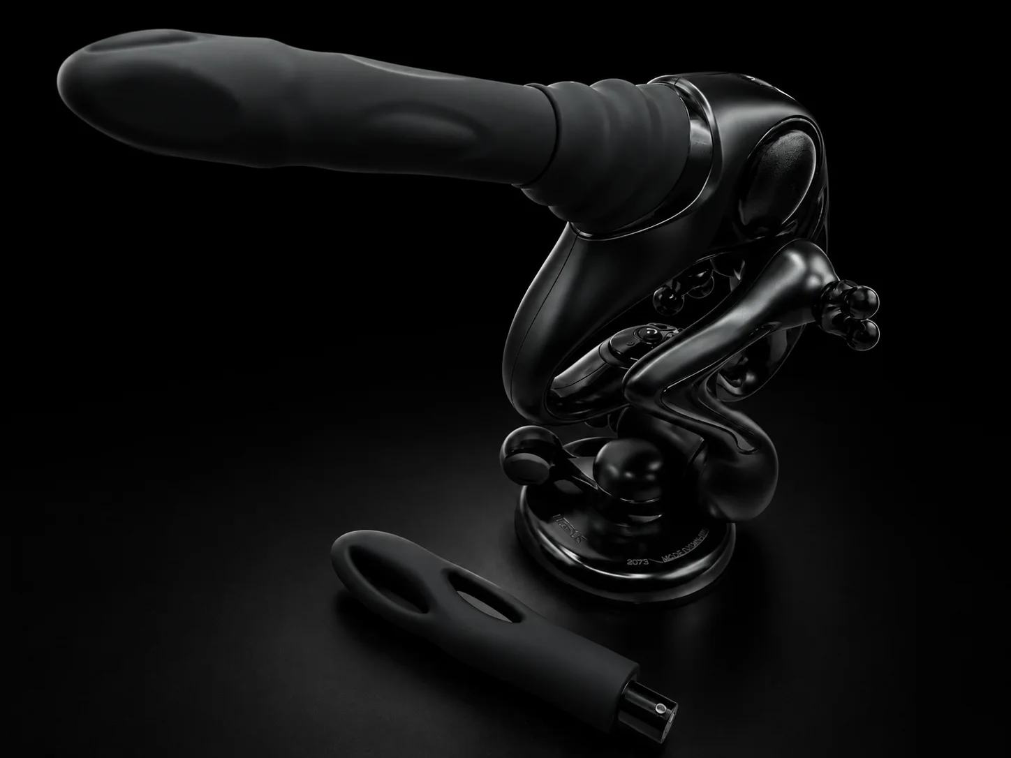UEROS Nexus sex machine on a black background: the glossy black main unit on its stand and lever-lock suction base with the thrusting attachment fitted, and the second attachment lying in front